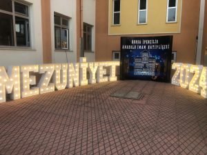 Bursa İpekçilik Anadolu İmam Hatip Lisesi 2024 mezuniyet töreni için Zeynepce Ajans tarafından kurulan ışıklı MEZUNİYET 2024 harfleri ve okul logosu içeren sahne dekorasyonu.